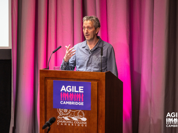 A speaker stands in front of pink curtains with an Agile Cambridge poster in front of them