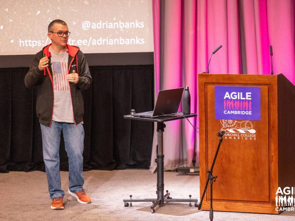 A person giving a presentation, they stand next to a podium with the Agile Cambridge logo on